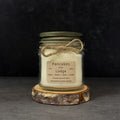 An 8 oz glass jar candle with off-white wax, an antique gold metal lid, and a rustic paper label reading 'Pancakes at the Lodge,' tied with twine and displayed on a wood slice against a dark background.