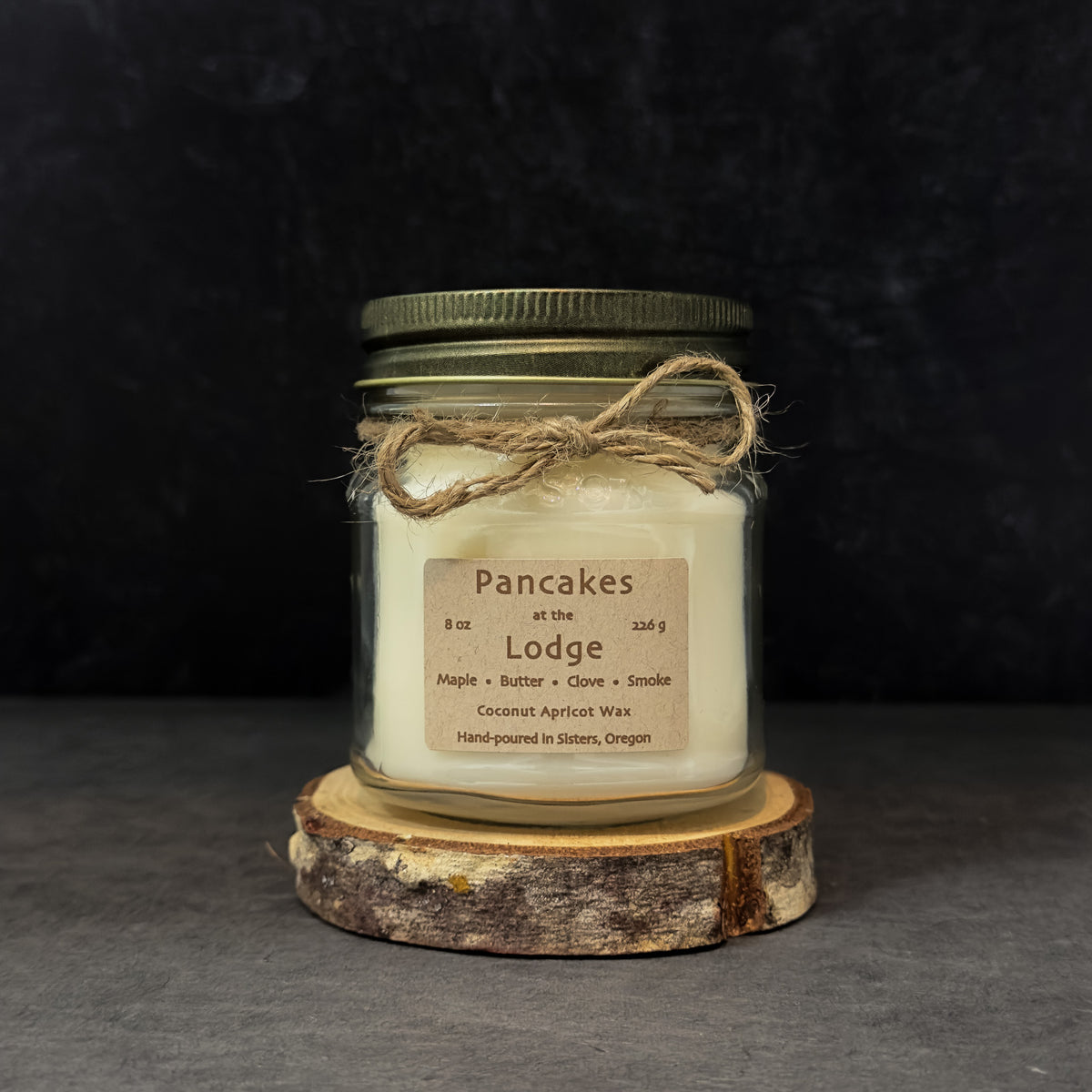 An 8 oz glass jar candle with off-white wax, an antique gold metal lid, and a rustic paper label reading 'Pancakes at the Lodge,' tied with twine and displayed on a wood slice against a dark background.
