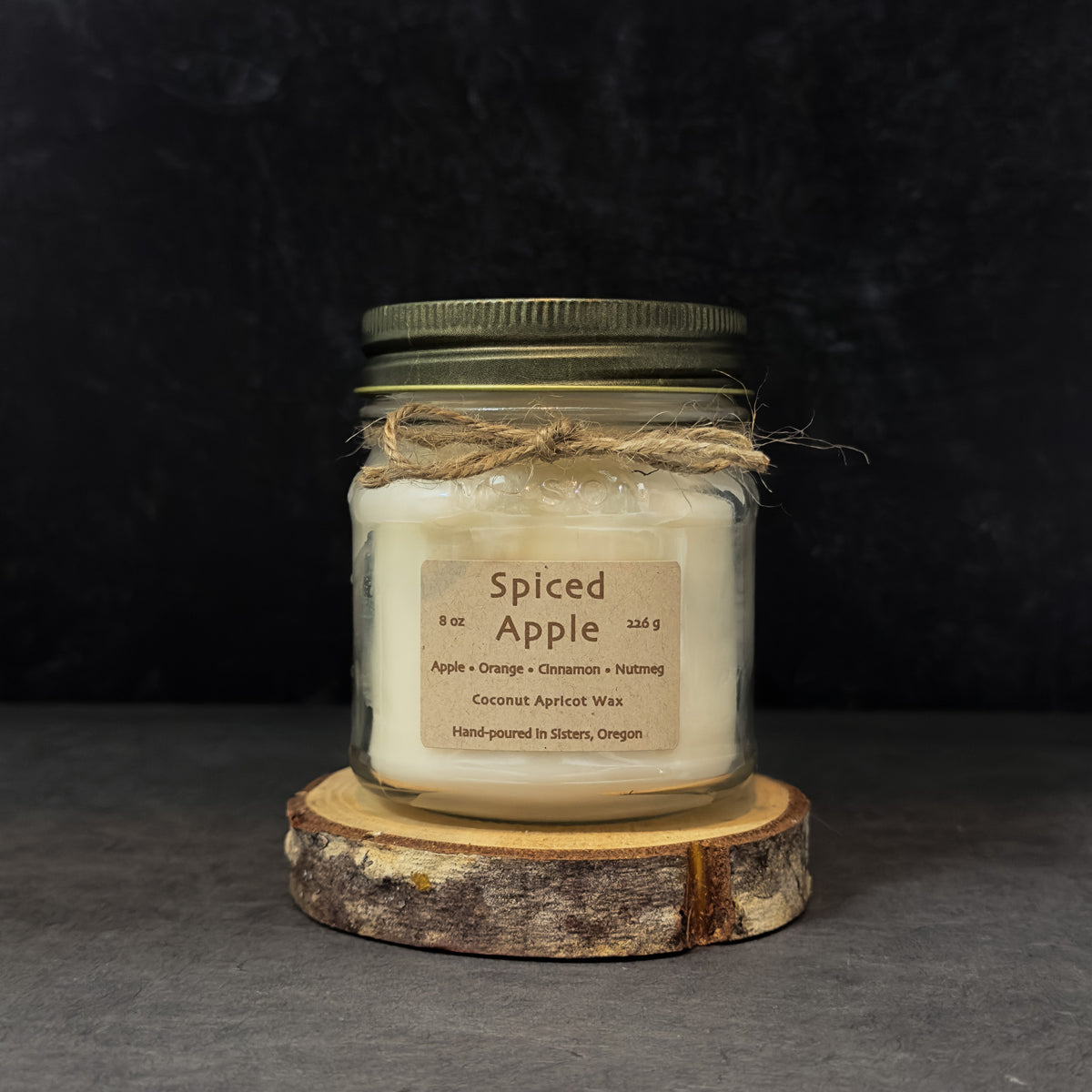 A glass jar candle with an antique gold metal lid and a rustic twine bow, labeled 'Spiced Apple' on a dark textured background, resting on a round wood slice coaster.
