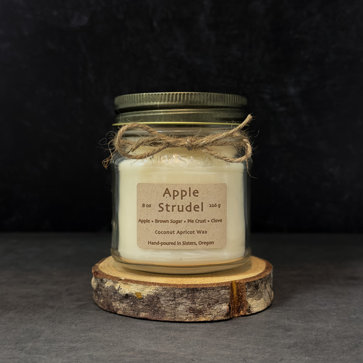 An 8 oz glass jar candle with cream-colored wax, an antique gold metal lid, and a brown paper label reading 'Apple Strudel.' The jar is wrapped with twine and sits on a round wood slice against a dark textured background.