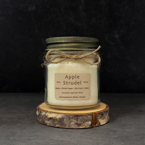 An 8 oz glass jar candle with cream-colored wax, an antique gold metal lid, and a brown paper label reading 'Apple Strudel.' The jar is wrapped with twine and sits on a round wood slice against a dark textured background.