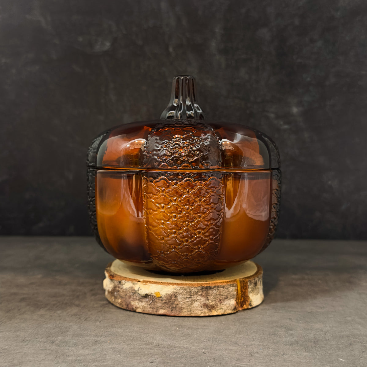 An amber glass pumpkin-shaped lidded box with embossed texture details, displayed on a rustic wood slice stand against a dark gray textured background.