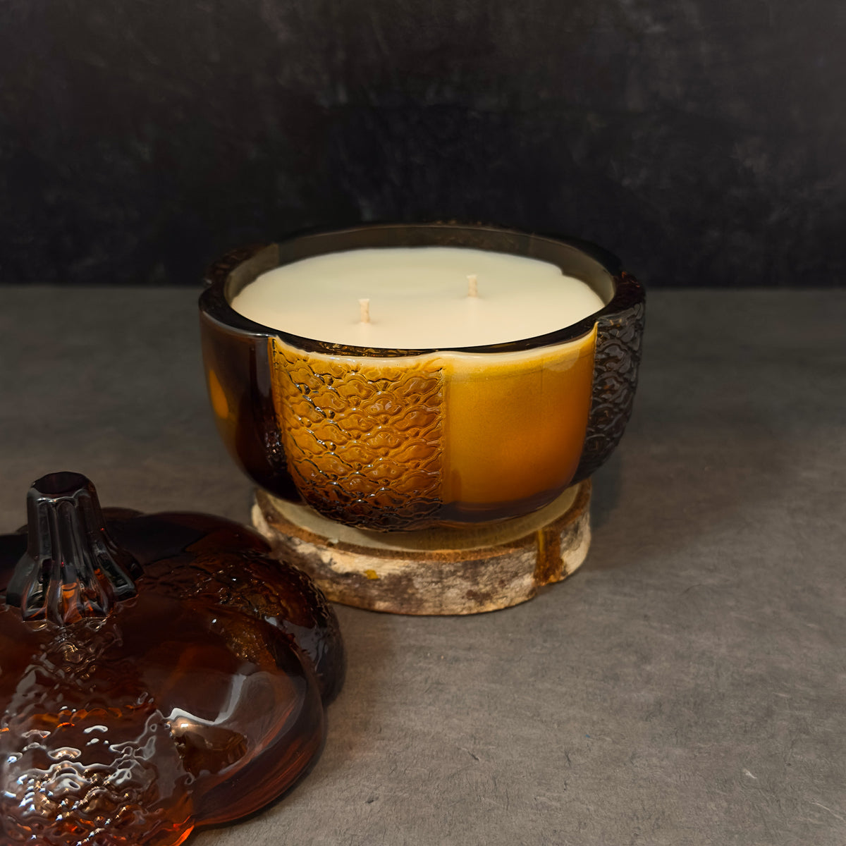 A two-wick candle in a textured amber glass bowl with a white wax fill, displayed on a rustic wood slice coaster next to its matching amber glass lid on a gray surface.