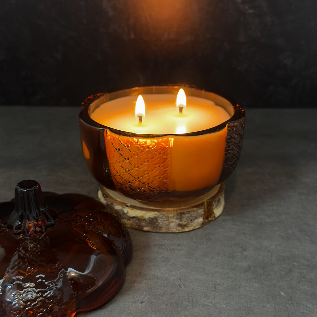 A lit amber glass candle holder with two flames, filled with warm yellow wax, set on a rustic wood slice base against a dark textured background. A matching amber glass lid is partially visible in the foreground.